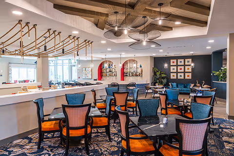 A spacious dining area with multiple tables and chairs featuring orange and blue upholstery. The room has a decorative ceiling with wooden beams and modern light fixtures. There is a bar area with shelves holding bottles and glasses, and a seating area with a blue couch and framed artwork on the wall.