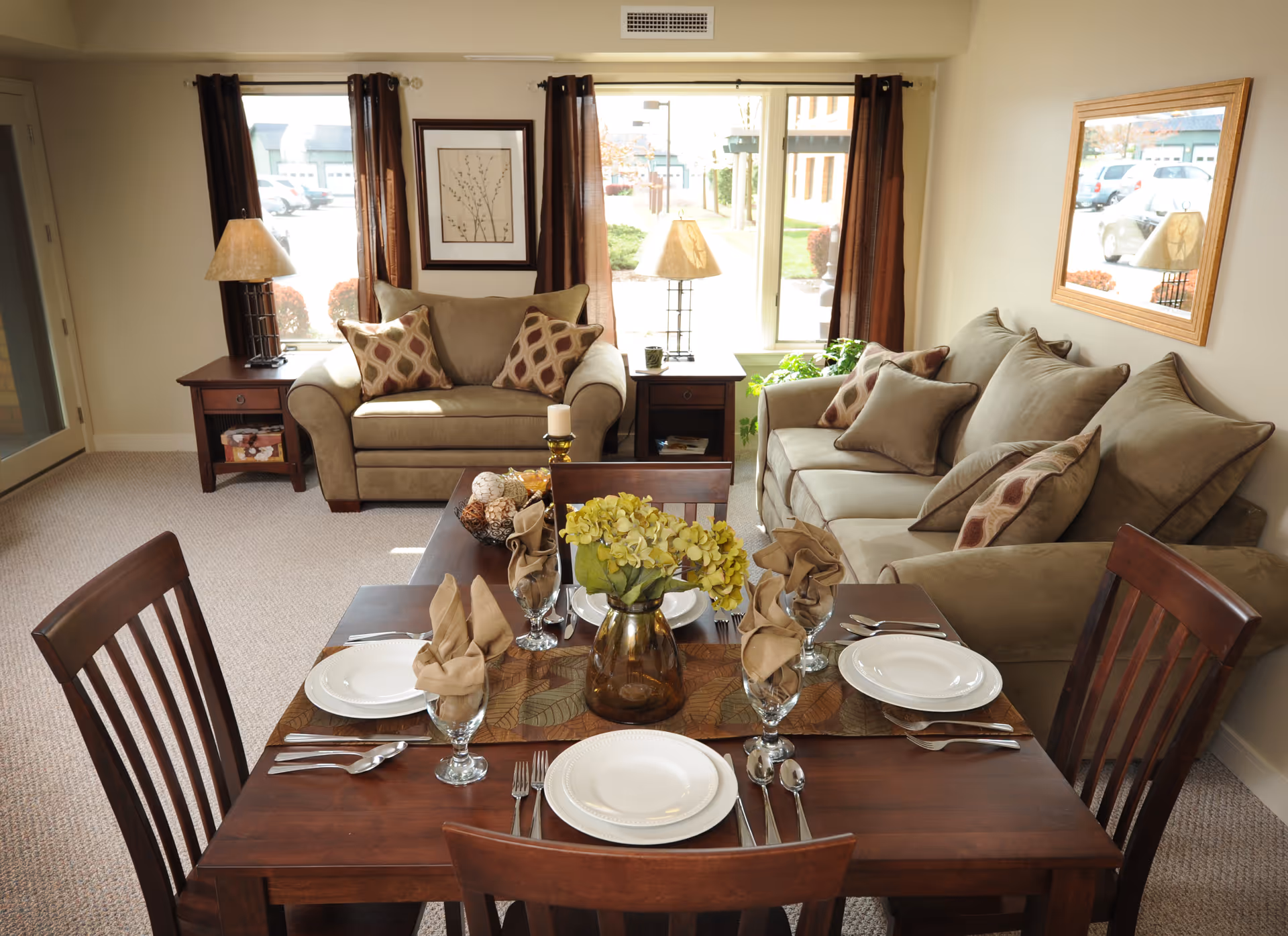 A cozy living and dining area featuring a wooden dining table set for four with plates, glasses, and beige napkins. Behind the table are two beige sofas with patterned throw pillows, two side tables with lamps, a framed picture on the wall, and a large window with brown curtains letting in natural light.