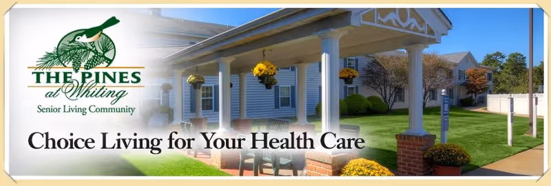 Exterior view of a senior living community named The Pines at Whiting, showing a covered porch with hanging flower baskets, a well-maintained lawn, and a building in the background under a clear blue sky.