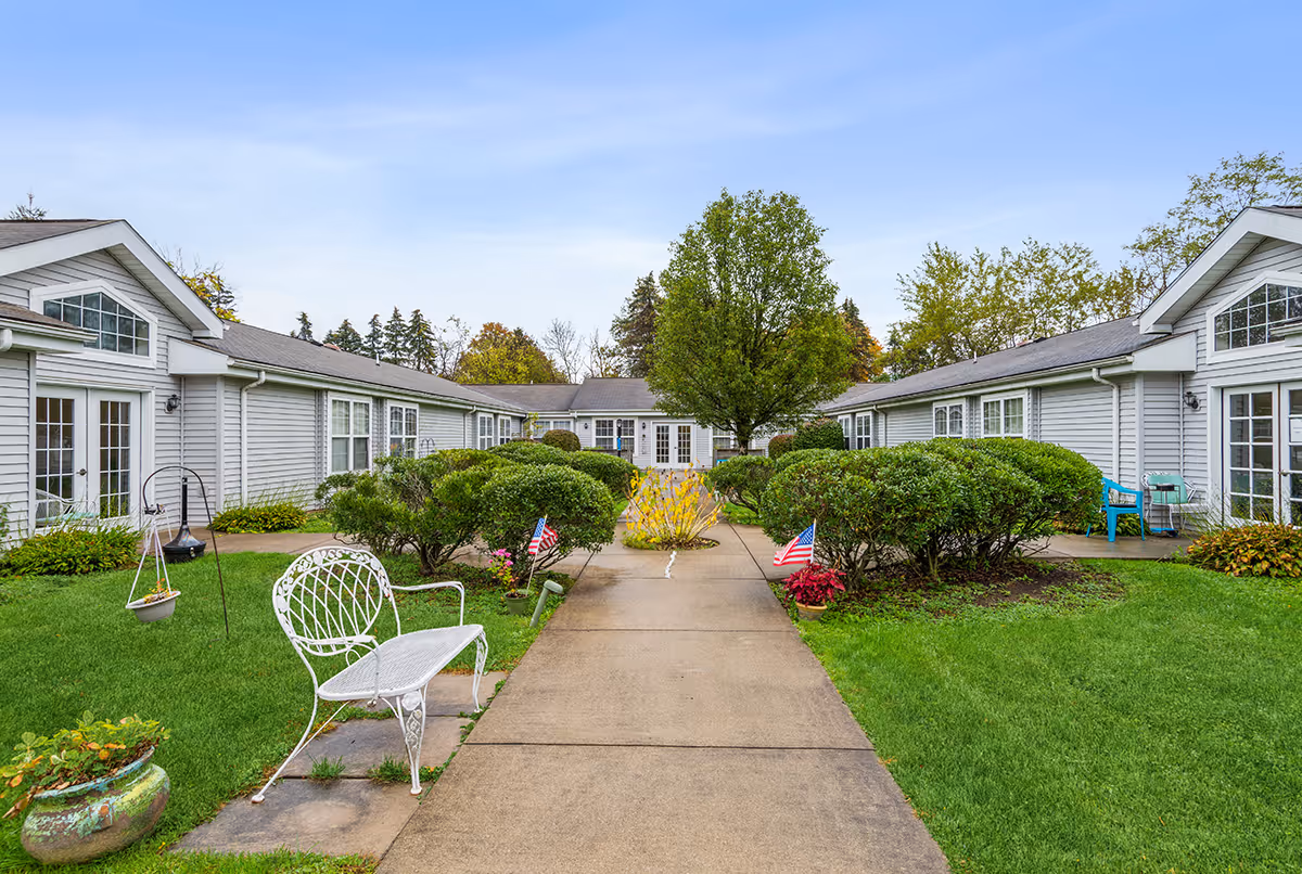 A central paved walkway through a landscaped courtyard flanked by single-story gray residential wings, benches, shrubs, and a tree.