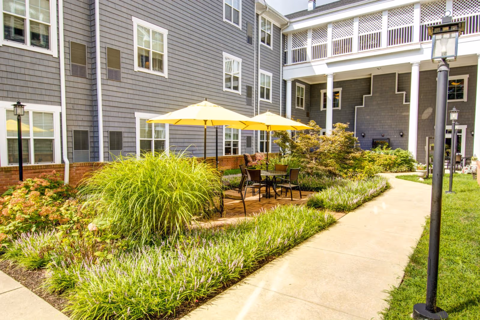 Courtyard patio at Symphony Manor with yellow umbrellas over tables and chairs, landscaped plantings and a walkway alongside the gray-sided building.