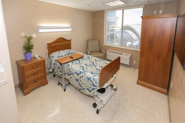 A small, clean bedroom in a care facility featuring a single hospital-style bed with a blue and white patterned blanket. Next to the bed is a wooden nightstand with a purple potted plant and a telephone. There is a beige armchair near a window with blinds, and a wooden wardrobe on the right side. The room has beige walls and a tiled floor.