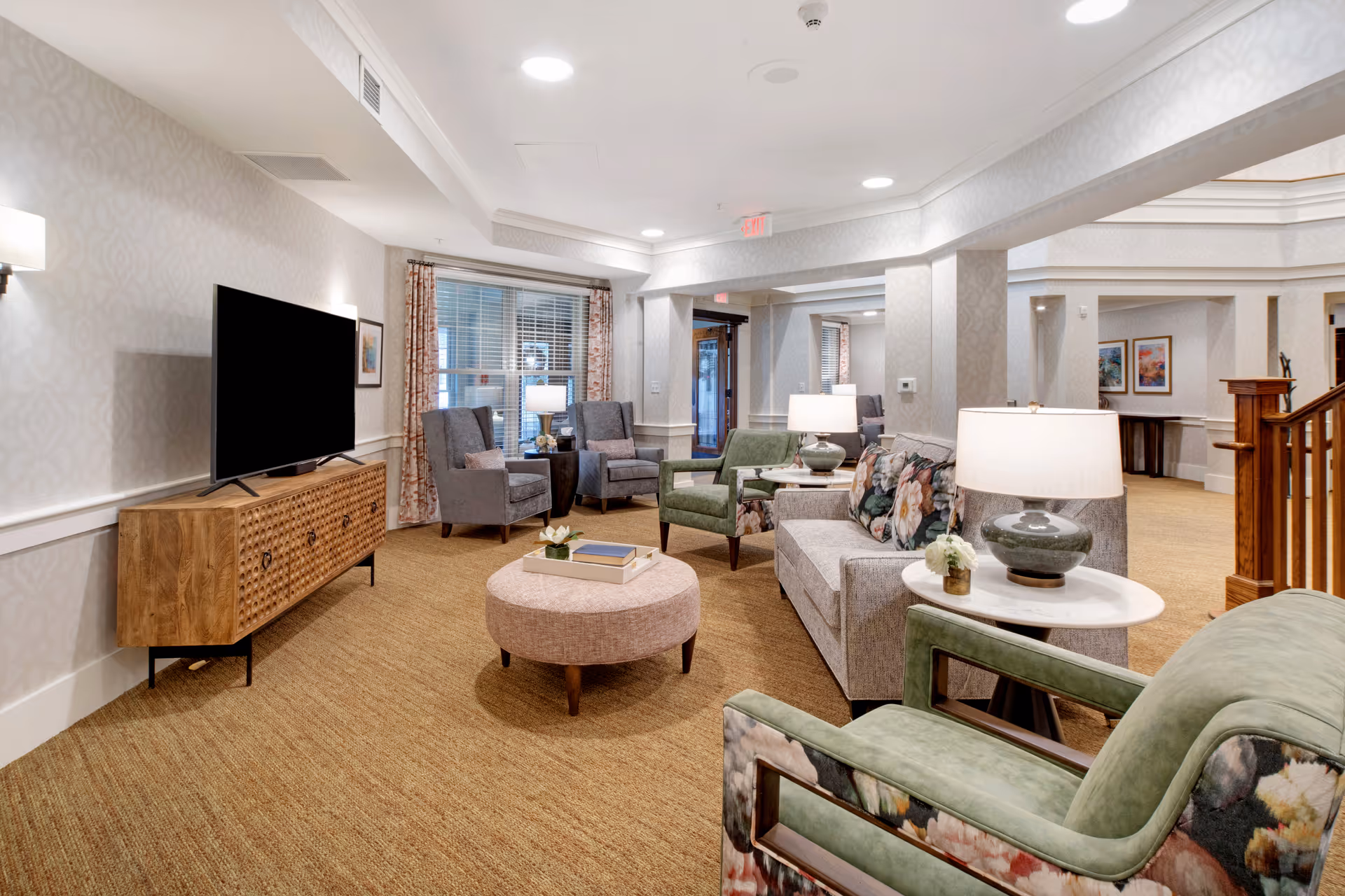 A well-lit senior living facility common area with a large flat-screen TV on a wooden console, several upholstered chairs and sofas with floral cushions, round ottoman, side tables with lamps, and framed artwork on the walls. The space has beige carpeting and light-colored wallpaper with a subtle pattern.