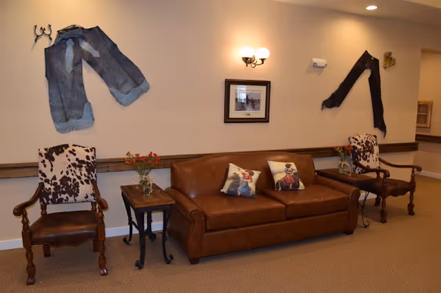 A cozy seating area with a brown leather sofa adorned with two decorative pillows featuring cowboy images. On each side of the sofa, there are wooden armchairs with cowhide upholstery. Two small wooden side tables hold vases with red flowers. The wall behind the seating area is decorated with a framed picture, a pair of denim chaps, and a denim jacket, all mounted as rustic decor. The lighting includes a wall-mounted double sconce with warm light.