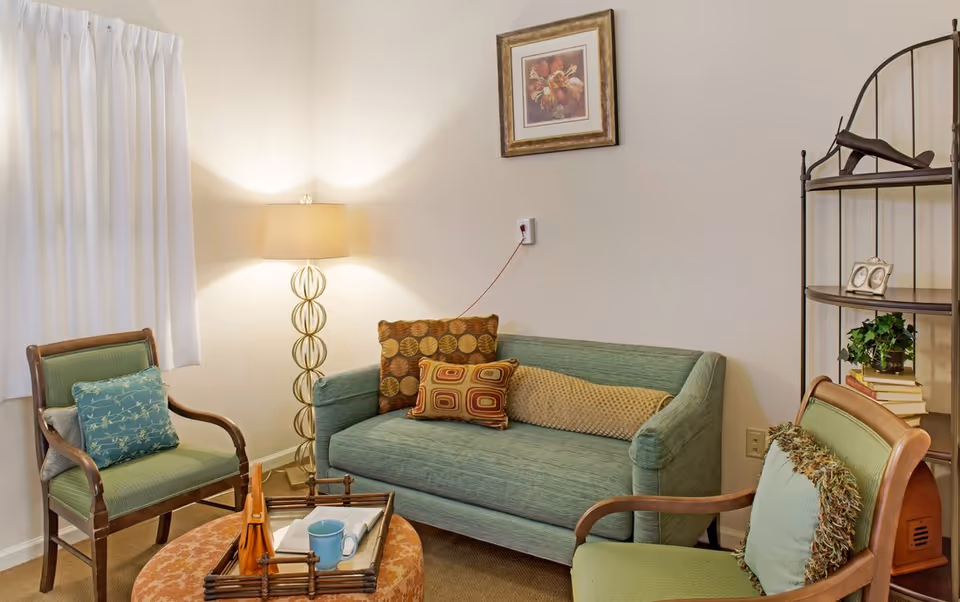 Cozy living room with a green loveseat and matching chairs around a small table, floor lamp, wall art, and a shelving unit.