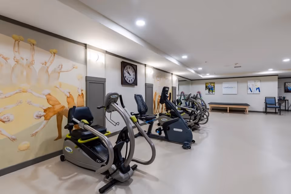 A fitness room with several exercise machines including recumbent bikes and elliptical trainers. The walls are decorated with artwork depicting people playing with balls. There is a large clock on the wall and some chairs and a bench along the far wall.