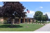 Single-story brick building with an American flag on a flagpole in front, surrounded by green grass, trees, and a sidewalk under a blue sky with some clouds.