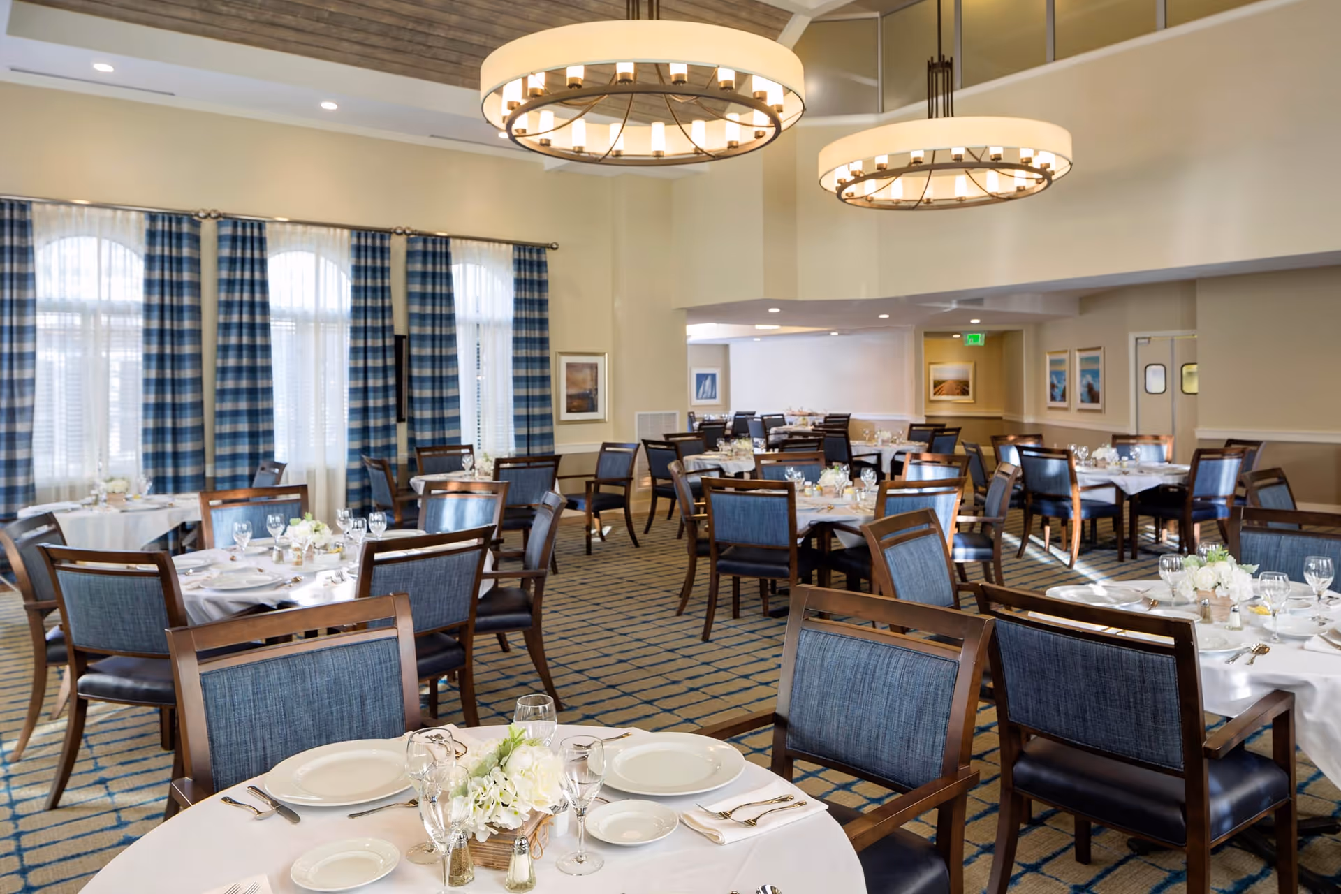 A spacious dining room with multiple round tables set with white tablecloths, plates, glasses, and floral centerpieces. The room features large windows with blue and white checkered curtains, modern circular chandeliers, and a patterned carpet.