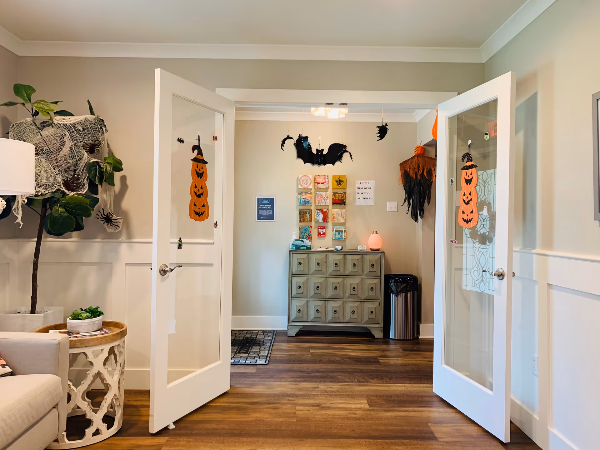 Open double glass doors lead into a bright, decorated common living area with a side table, potted plant, and a small cabinet adorned with Halloween decorations.