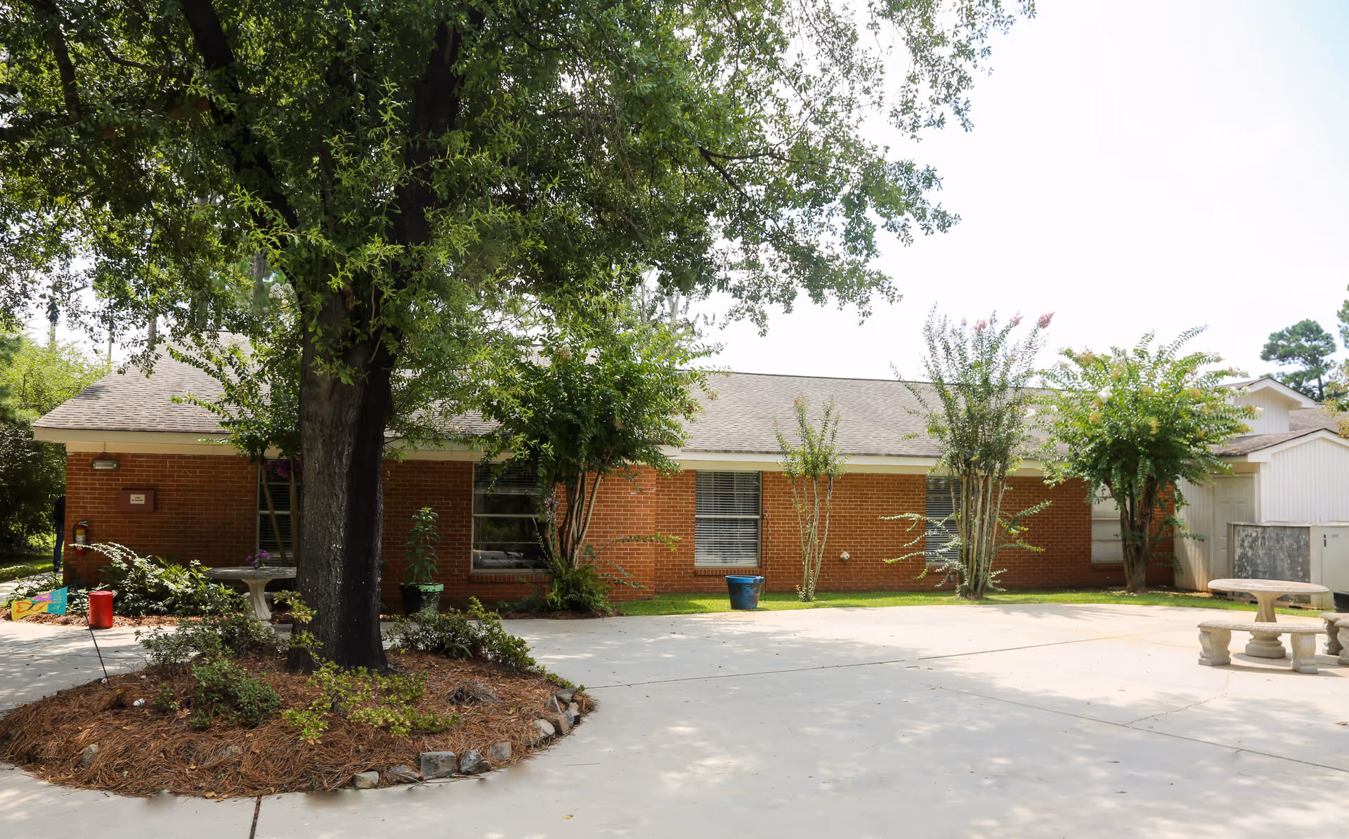 Front view of a single-story brick building with a large tree, landscaping, and a paved courtyard with a stone picnic table.