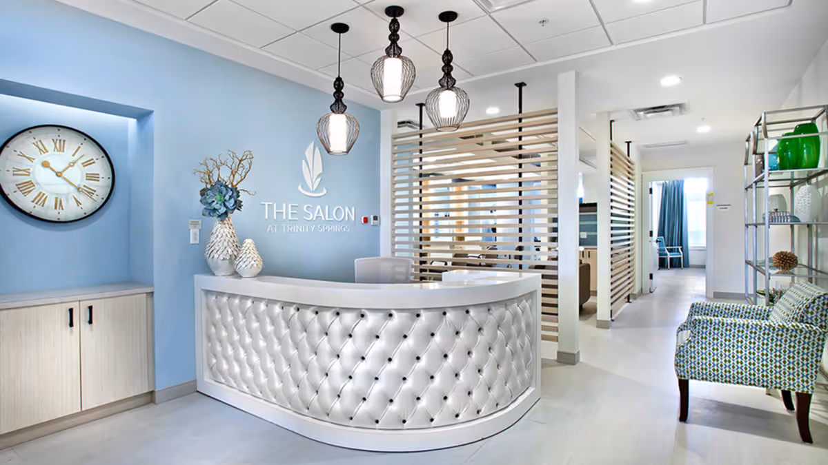 Reception area of 'The Salon at Trinity Springs' featuring a tufted white curved desk, blue walls, pendant lights, and patterned seating.