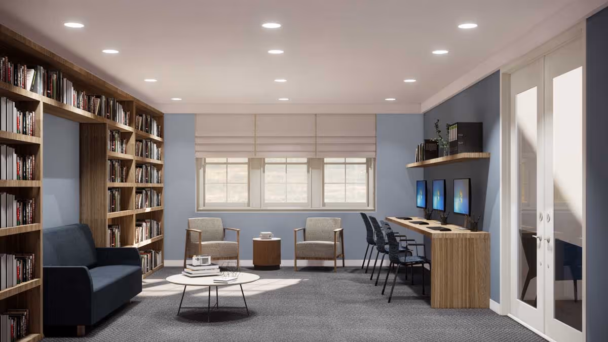 A well-lit room with blue walls featuring a large wooden bookshelf filled with books on the left side, a dark blue sofa, two armchairs with a small round table between them near the window, and a row of four computer workstations with chairs along the right wall. The floor is carpeted in gray, and there are recessed ceiling lights. A set of double glass doors is visible on the right side.
