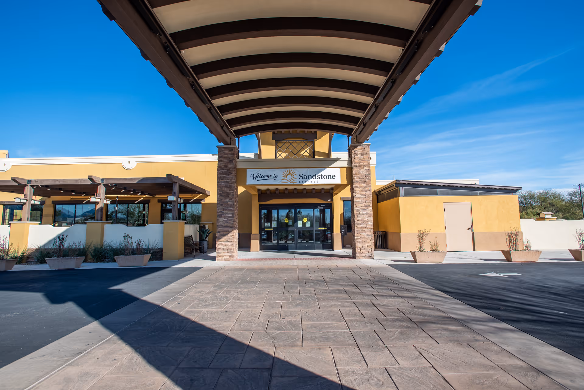 Front exterior view of Sandstone Estates facility with a covered entrance supported by stone pillars, yellow and beige walls, and a clear blue sky.