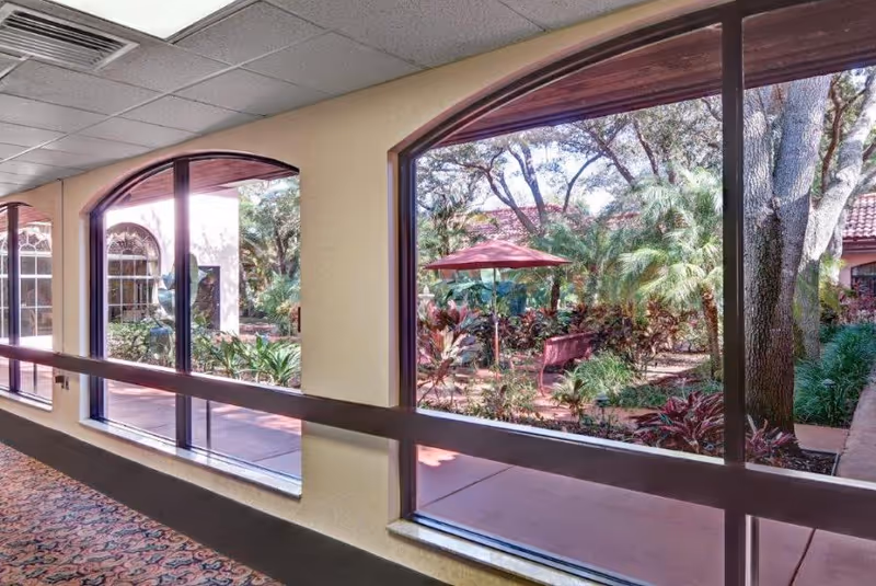 View through large arched windows from an indoor hallway showing a landscaped outdoor garden area with trees, plants, a bench, and a red umbrella.