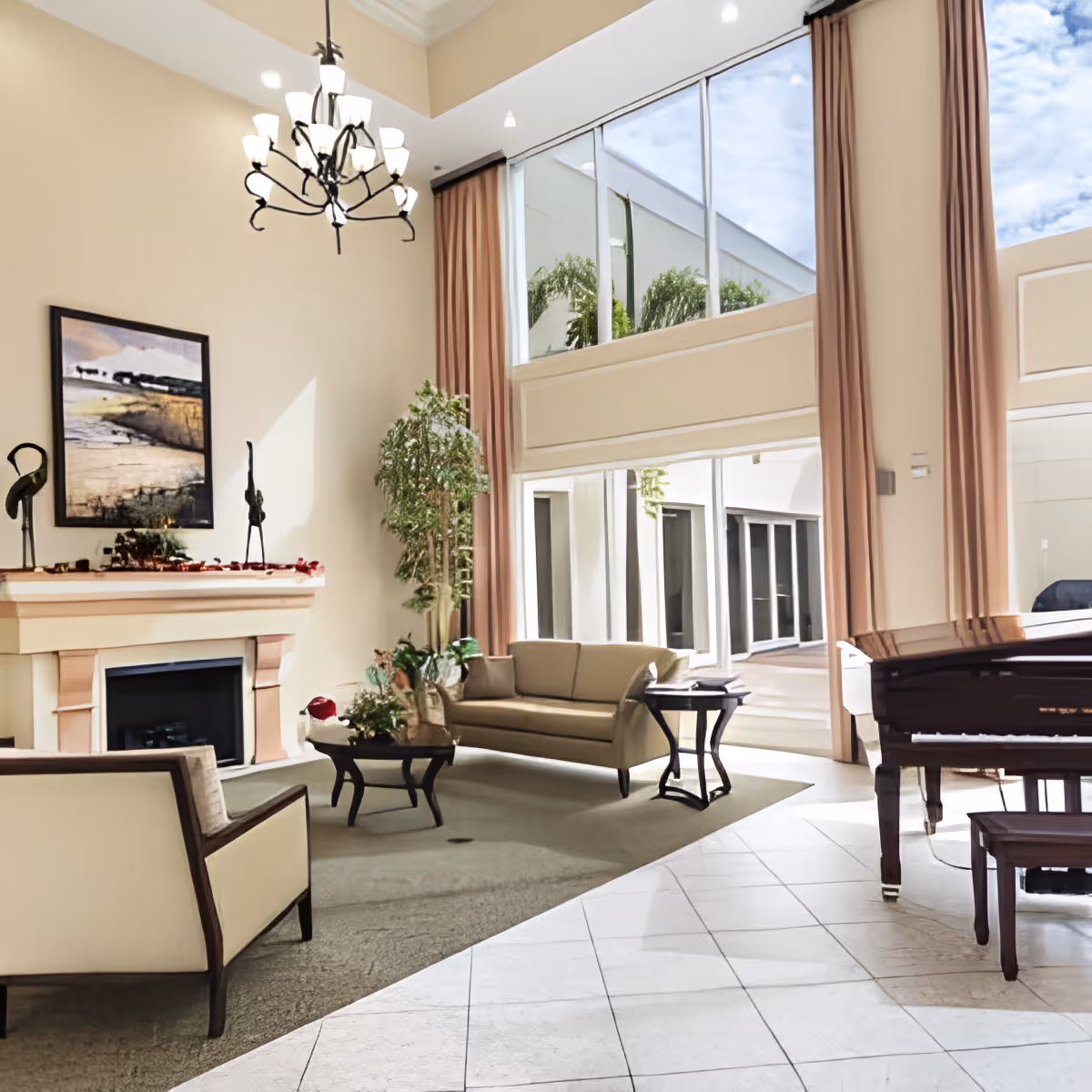 A bright and spacious living room area with high ceilings and large windows letting in natural light. The room features a beige sofa, armchair, coffee table, side table, a grand piano, and a fireplace with decorative items on the mantel. There are tall curtains framing the windows and a chandelier hanging from the ceiling.