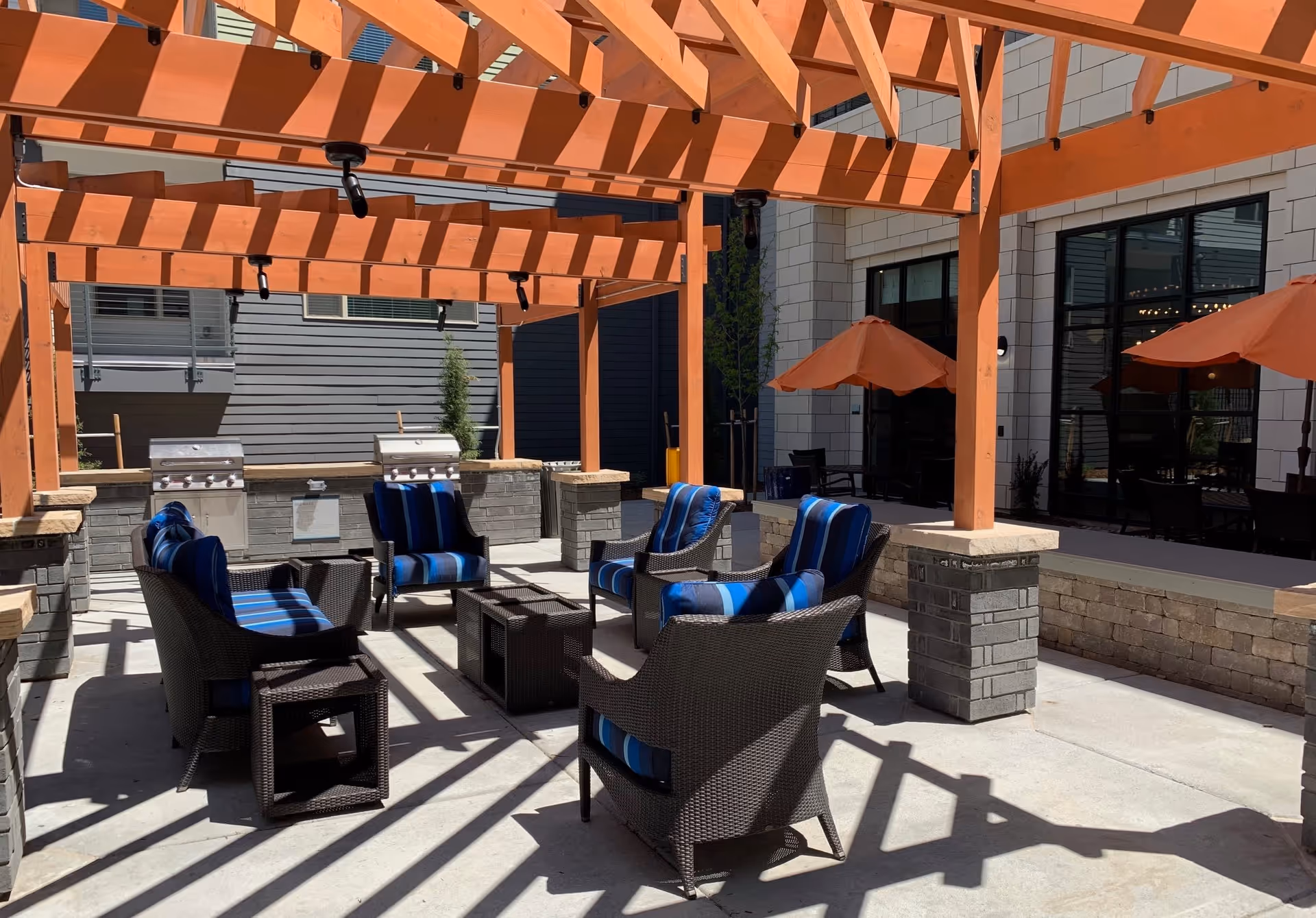 Shaded outdoor patio with a wooden pergola, wicker lounge chairs with blue-striped cushions around a low table, gas grills, and orange umbrellas by the building.