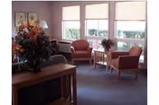 A cozy living room area with two armchairs and a small wooden table between them, decorated with a flower arrangement. There is a wooden console table with a large floral arrangement in the foreground, and three windows with blinds letting in natural light.