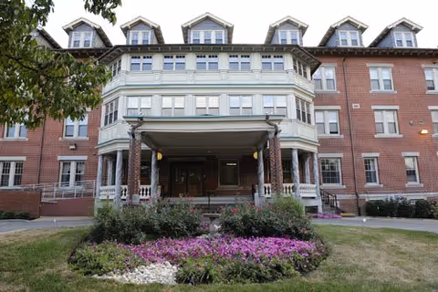 Brick senior living building with a covered entry and colorful front flower beds.
