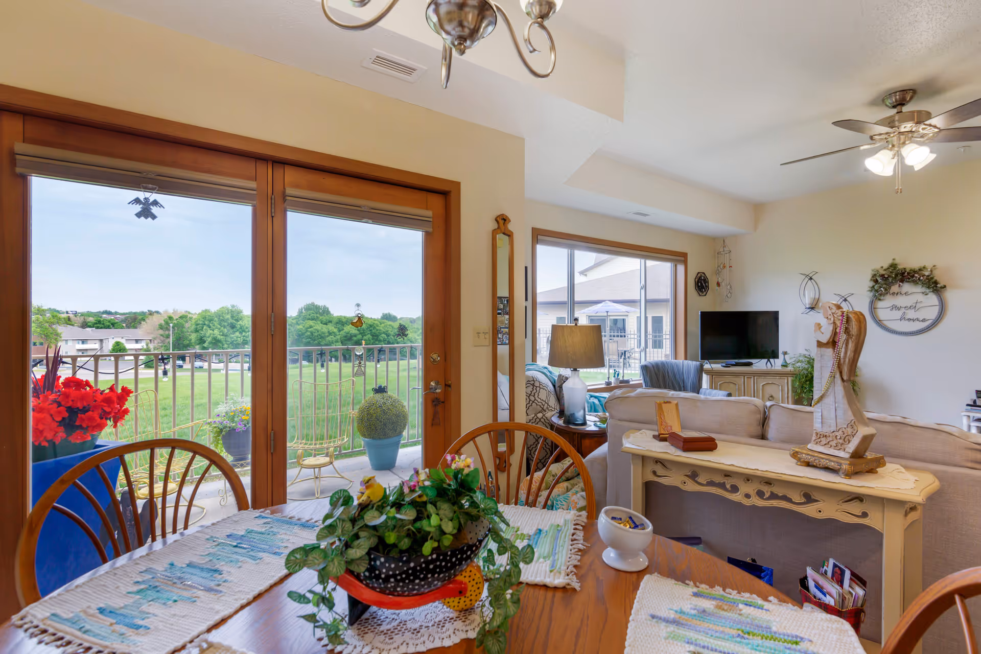View of a cozy senior living dining and living room area with a wooden dining table decorated with placemats and a potted plant. The room has large glass doors and windows that open to a balcony with outdoor plants and a green lawn beyond. The living room features a beige sofa, a TV on a wooden stand, a ceiling fan, and decorative items including a lamp and wall art.