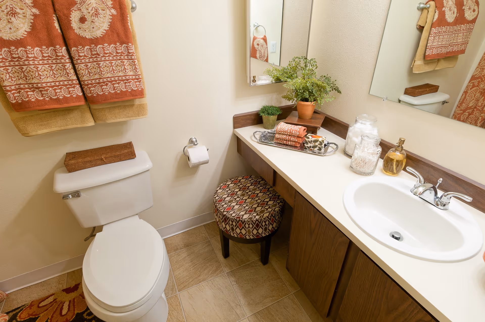A clean bathroom featuring a white toilet with a brown tissue box on top, a toilet paper holder with a roll of toilet paper, a countertop with a white sink and silver faucet, decorative jars, a soap dispenser, a small potted plant, folded towels on a tray, and a patterned cushioned stool underneath the counter. Two orange and beige patterned towels hang on the wall above the toilet, and a large mirror is mounted above the sink.