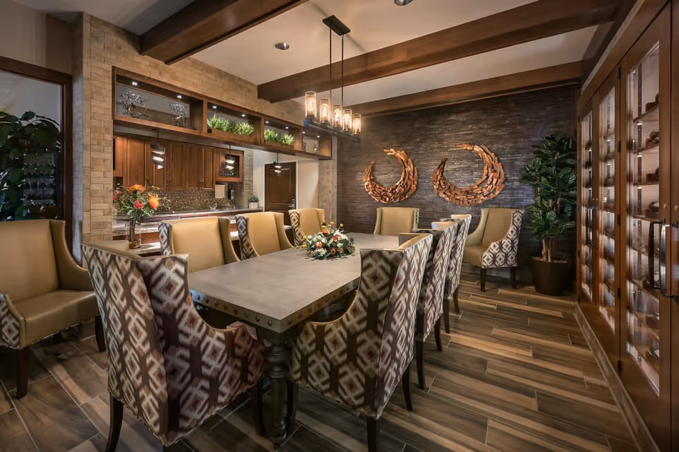 A stylish communal dining room with a long table surrounded by patterned upholstered chairs, warm wood beams, decorative wall art and cabinetry.