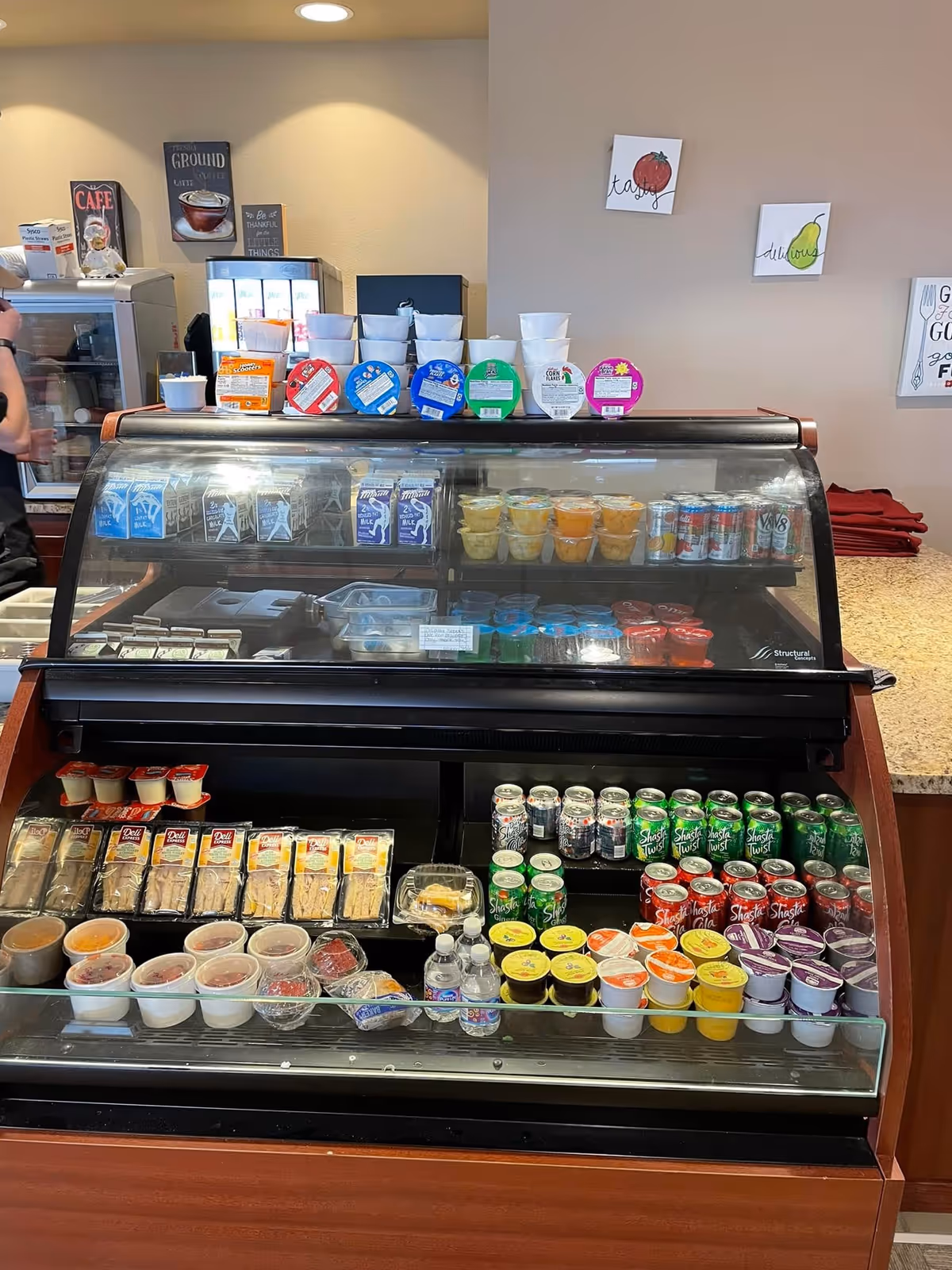 A refrigerated display case filled with various packaged food and drink items including milk cartons, fruit cups, canned beverages, pudding cups, sandwiches, and bottled water. The background shows a beige wall with small decorative signs featuring fruit illustrations and motivational quotes.