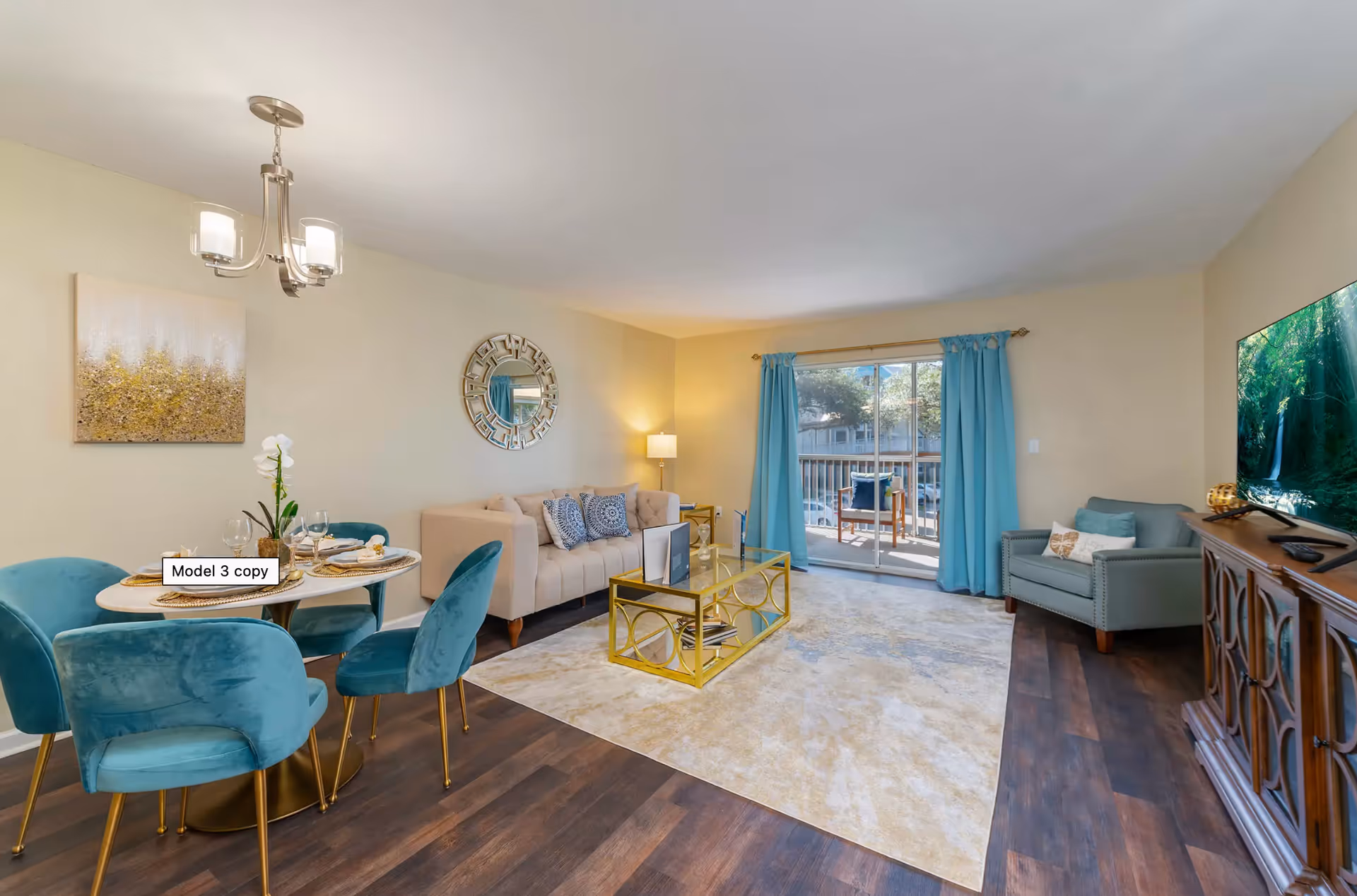 A bright and modern living room and dining area with a beige sofa adorned with blue patterned pillows, a glass coffee table with a gold frame, a large area rug, and a flat-screen TV on a wooden cabinet. The dining area features a round table with four teal velvet chairs and a chandelier overhead. Blue curtains frame a sliding glass door leading to a balcony with outdoor seating.