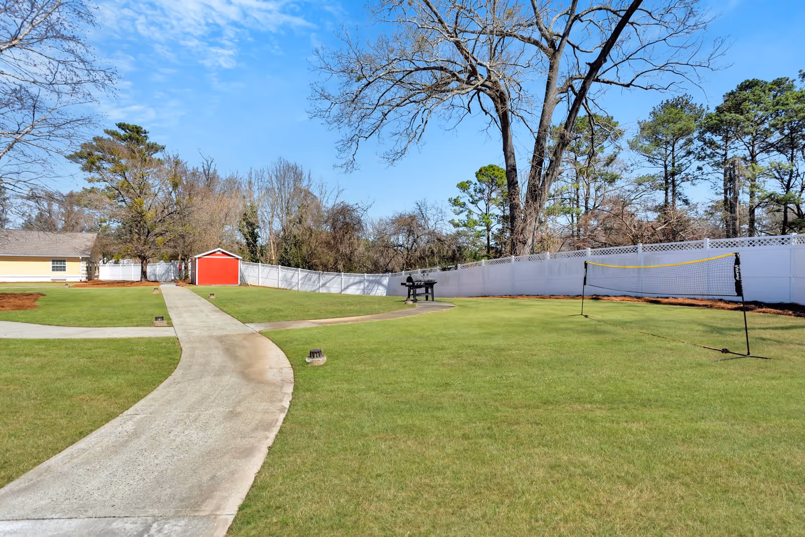 A spacious outdoor area with a curved concrete pathway leading to a small red shed. The area is surrounded by a white fence and tall trees. There is a portable badminton net set up on the grass and a barbecue grill near the fence.