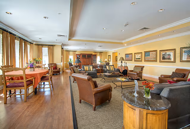 A spacious, well-lit living room and dining area in a senior living facility. The living room features comfortable leather and fabric armchairs arranged around a coffee table on a large area rug. The walls are decorated with framed artwork, including American flags. The dining area has wooden tables and chairs with orange tablecloths and floral centerpieces. Large windows with curtains allow natural light to fill the room.