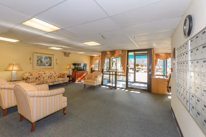 Cozy senior living lobby with floral sofa, armchairs, lamps, and glass entry doors.