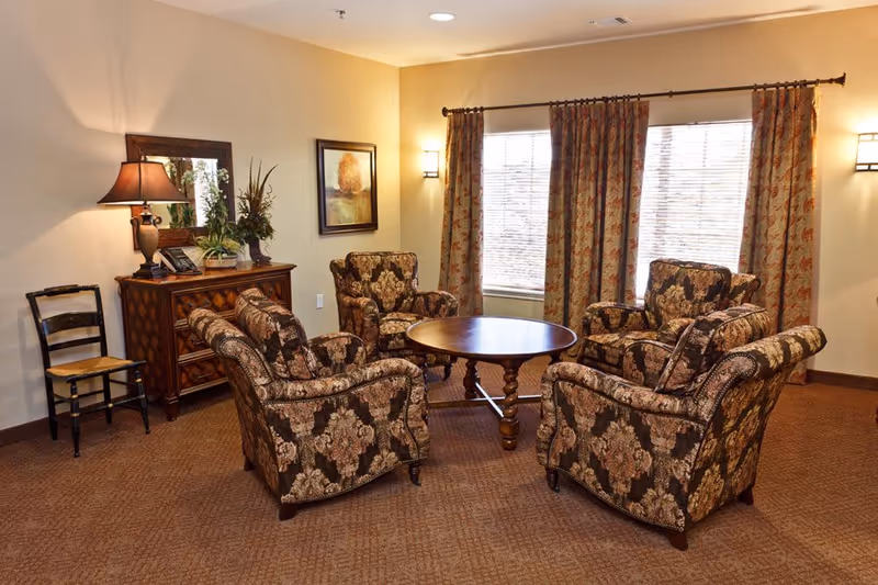 Cozy sitting area with four patterned armchairs arranged around a round wooden table in a warmly lit room with curtains and a side table.