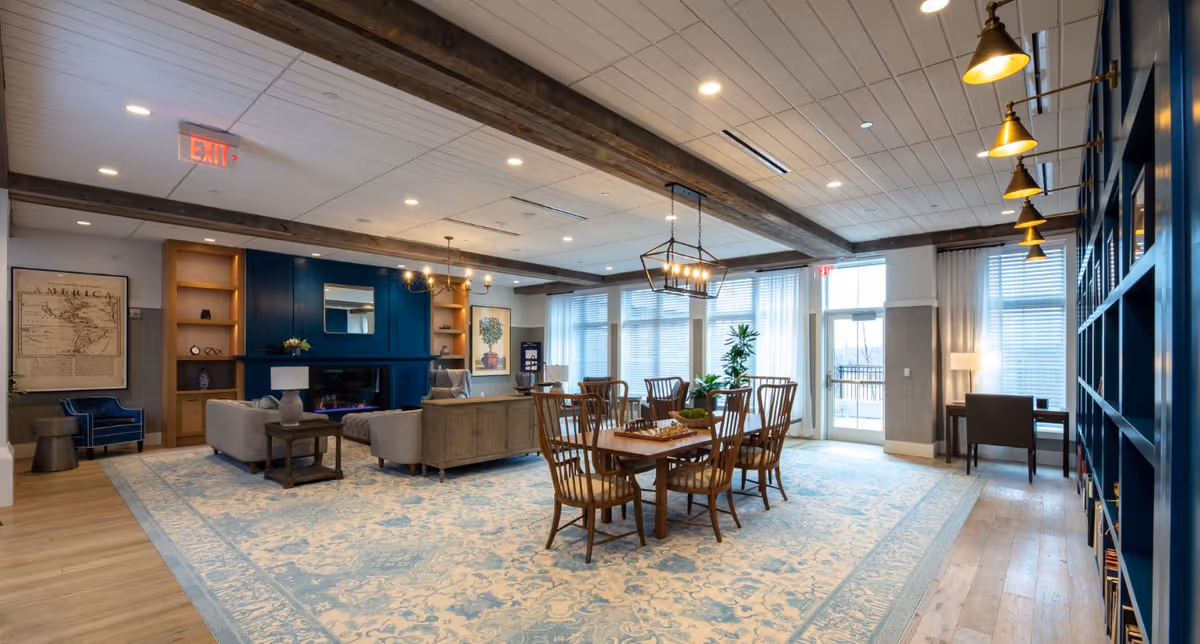 A spacious and well-lit common area in a senior living facility featuring a large patterned area rug, wooden dining table with chairs, comfortable sofas around a fireplace with a blue accent wall, bookshelves, and large windows allowing natural light to fill the room.