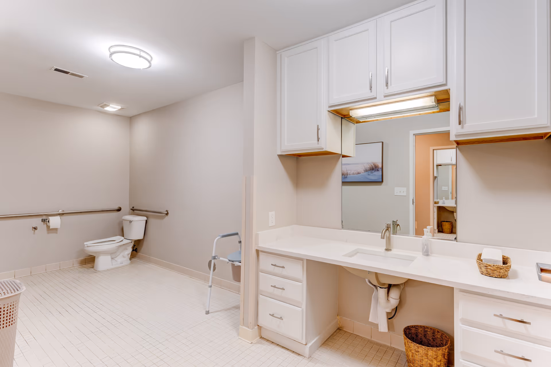 A spacious accessible bathroom in a senior living facility with a toilet, grab bars along the wall, and a long vanity with sink and cabinets.