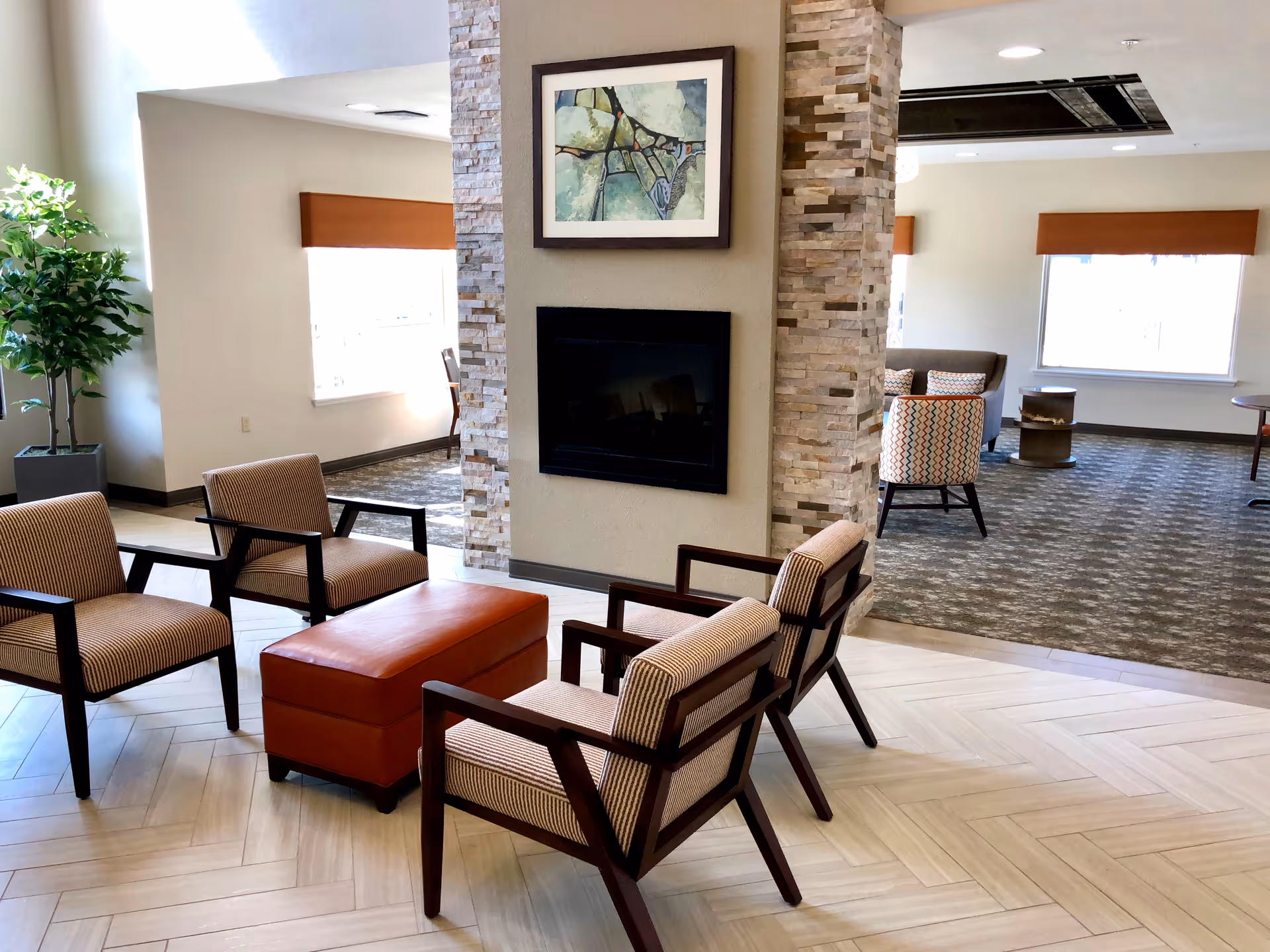A modern lounge area with four striped armchairs arranged around a rectangular orange ottoman. The space features a stone accent wall with a mounted flat-screen TV and a framed abstract painting above it. Large windows with orange valances allow natural light to fill the room, and a potted plant is visible in the corner.