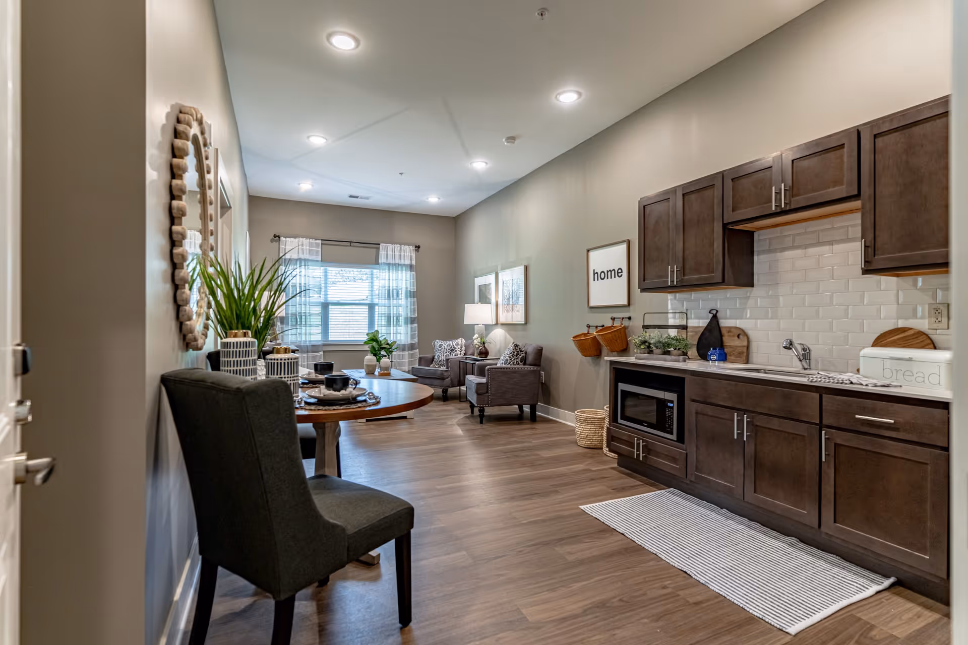 Interior view of a senior living facility apartment featuring a small kitchen area with dark wood cabinets, a microwave, and a white subway tile backsplash. A striped rug lies on the wooden floor in front of the kitchen. Adjacent to the kitchen is a round dining table with a dark upholstered chair and decorative plants. In the background, there is a living area with two armchairs, a side table with a lamp, framed artwork on the wall, and a window with sheer curtains letting in natural light.