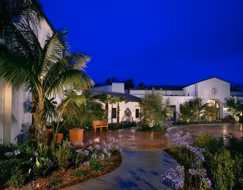Exterior view of a building at dusk with a clear deep blue sky. The building has white walls and an arched entrance, surrounded by well-maintained landscaping including palm trees, flowering plants, and shrubs. A paved walkway leads to the entrance.