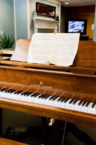 Close-up view of a wooden Young Chang piano with sheet music placed on the music stand. In the background, there is a fireplace with a mirror above it and a television mounted on the wall.