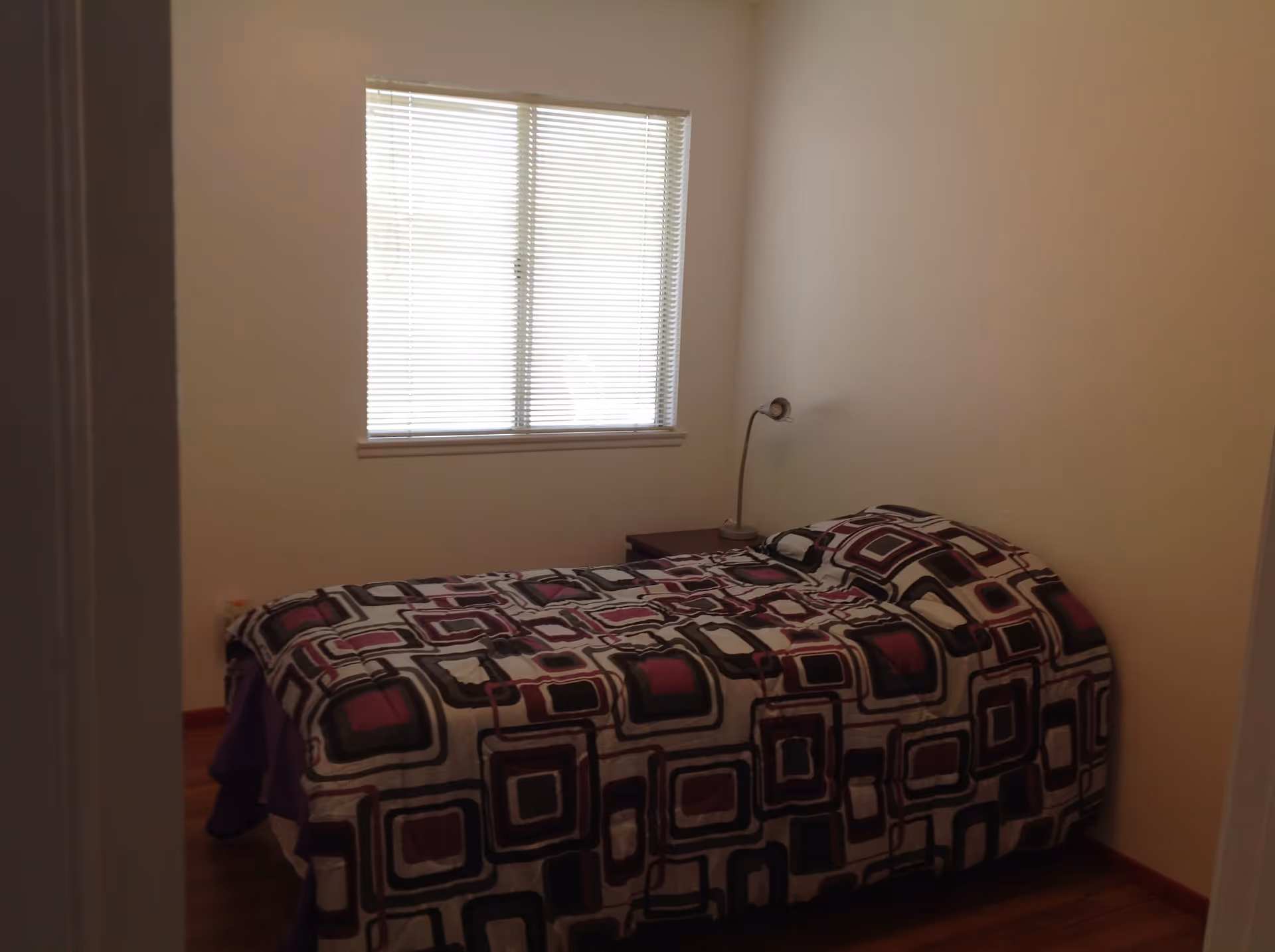 Small bedroom with a single bed covered in geometric-patterned bedding, a bedside table and lamp beneath a window with blinds.