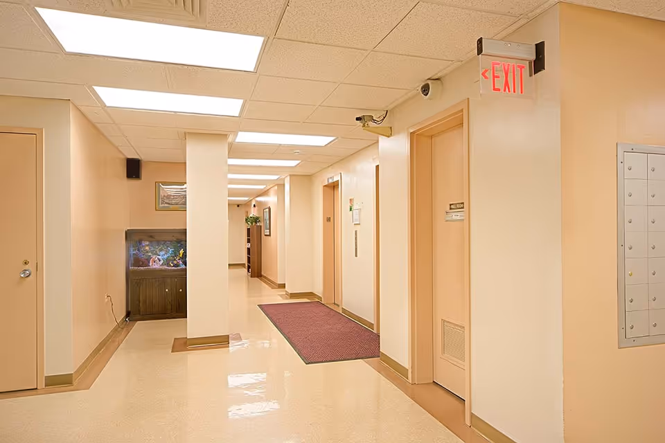 Well-lit interior hallway of a senior living facility with elevator doors, an EXIT sign, a fish tank, and a maroon runner rug.