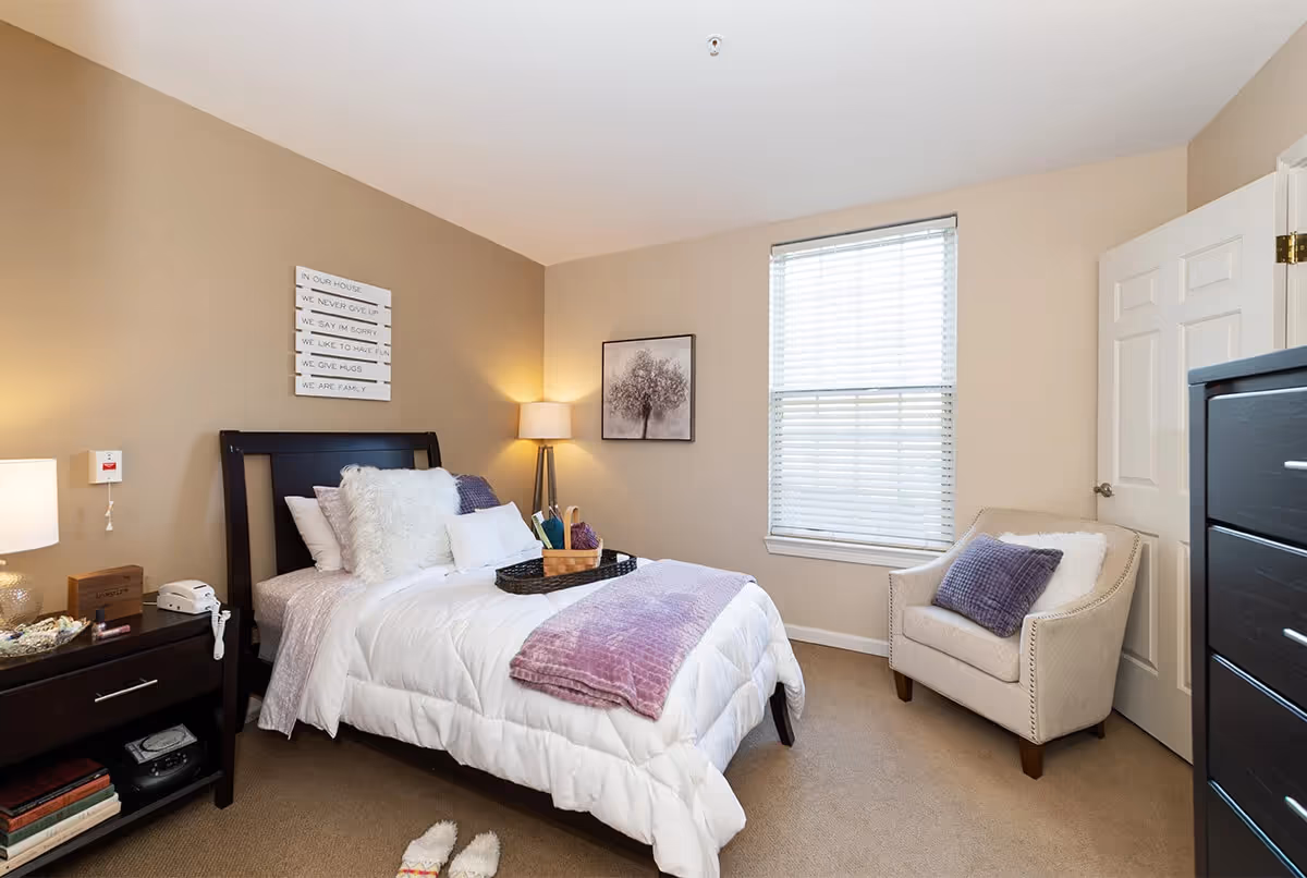 A cozy bedroom with a neatly made bed featuring white bedding and purple accents, a bedside table with a lamp and telephone, a beige armchair with a purple pillow, a window with blinds, and wall art including a framed tree picture and a sign with inspirational text.