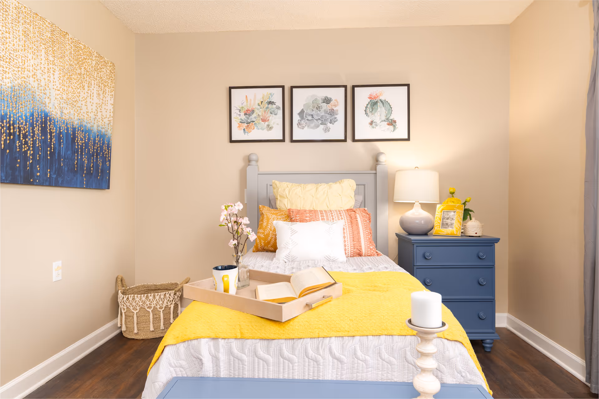 A cozy bedroom with a single bed featuring a gray headboard, white and yellow bedding, and multiple decorative pillows. A wooden tray with a book, a cup, and a small vase with flowers is placed on the bed. To the right of the bed is a blue nightstand with a lamp, a yellow picture frame, and a small plant. Three framed botanical prints hang on the beige wall above the bed. A woven basket is on the floor to the left, and a large abstract painting with gold and blue tones is on the left wall.