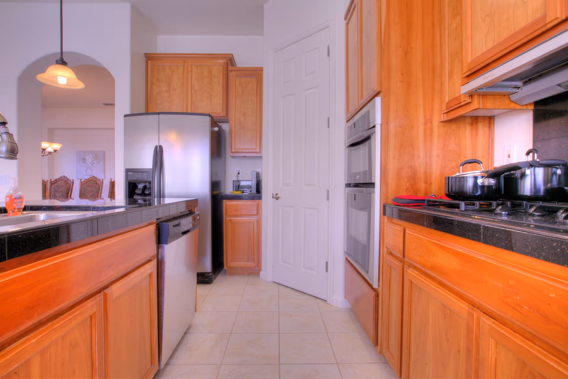 A modern kitchen with wooden cabinets, black granite countertops, a stainless steel refrigerator, dishwasher, double oven, and a gas stove with pots on it. There is a pendant light hanging from the ceiling and a glimpse of a dining area with chairs in the background.