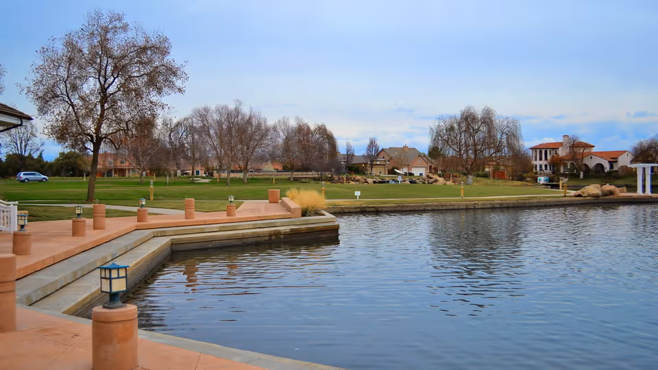 A lakeside promenade with terracotta walkways, small lanterns, grassy lawns and houses across the water.
