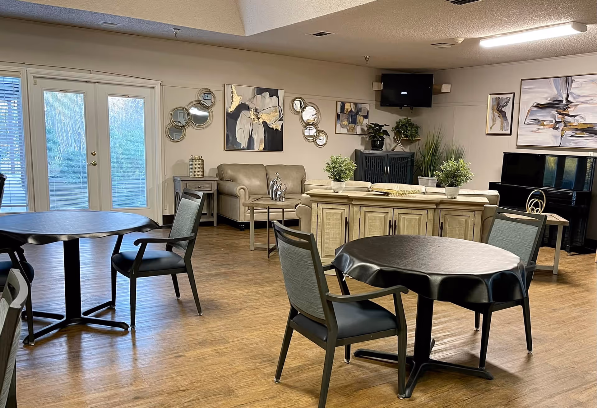A cozy common area in a senior living facility featuring round tables with chairs, a beige sofa, decorative plants, abstract wall art, a piano, and a wall-mounted television. French doors with blinds lead to an outdoor space.