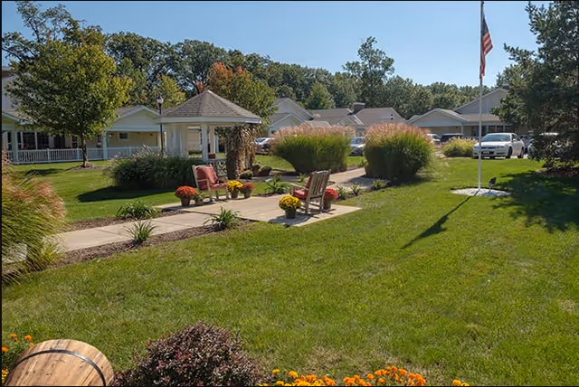 A well-maintained outdoor garden area at a senior living facility with green grass, a paved walkway, wooden chairs, potted flowers, a small gazebo, and several trees. Residential buildings and parked cars are visible in the background under a clear blue sky.