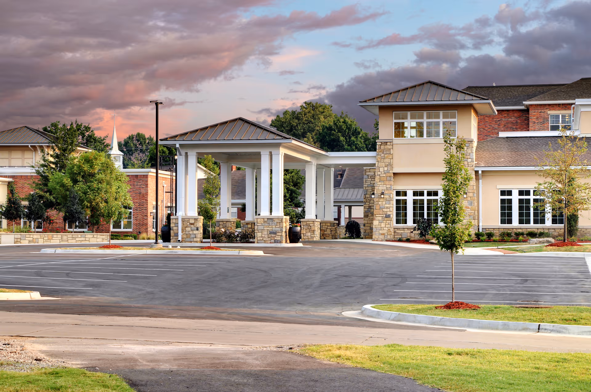 Front exterior of a senior living facility with a covered porte-cochère, empty parking lot, and landscaped grounds beneath a dramatic sky.
