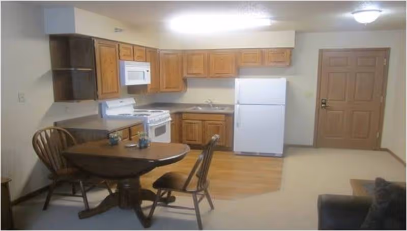 Interior view of a small kitchen and dining area in a senior living facility. The kitchen features wooden cabinets, a white refrigerator, a white stove with an overhead microwave, and a sink. In front of the kitchen is a wooden dining table with two chairs. The room has a mix of carpet and wood flooring, and a closed wooden door is visible in the background.
