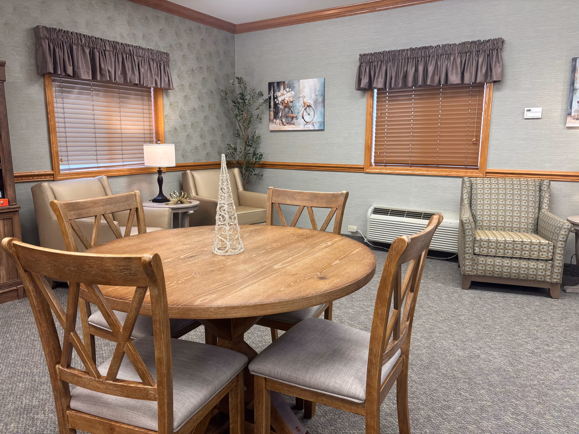 Round wooden dining table with four chairs in a cozy common room featuring armchairs, a lamp, wall art and window blinds.