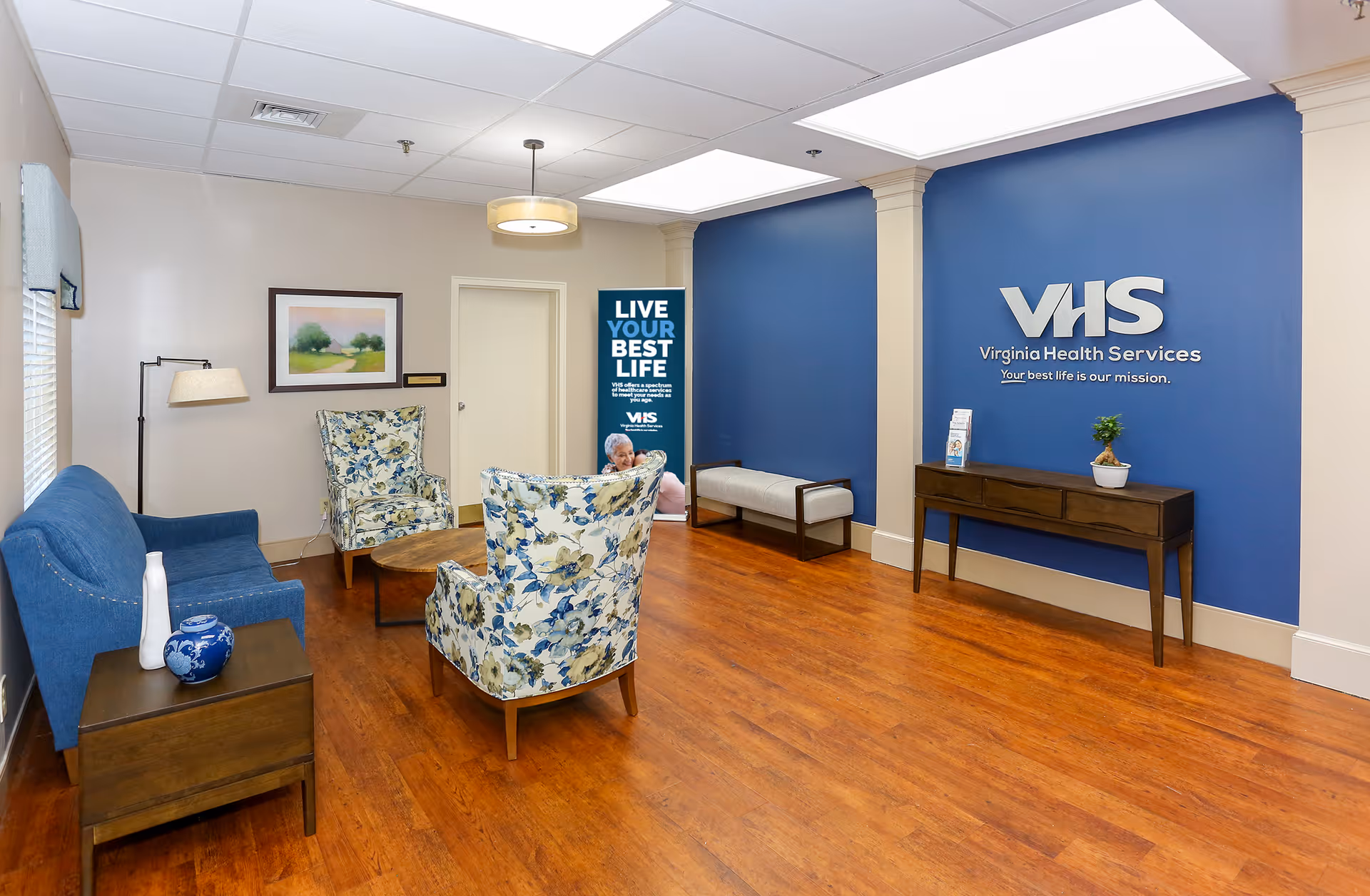 Bright waiting area with a blue accent wall displaying "VHS Virginia Health Services", wooden floors, a blue sofa, two floral armchairs, and a small console table.