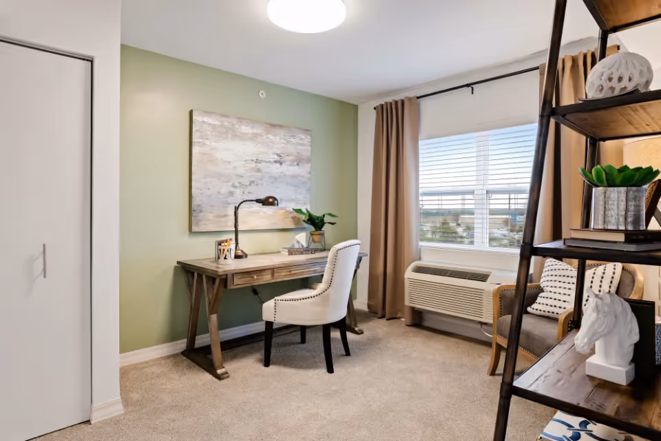 Furnished living area with a wooden desk and upholstered chair against a green accent wall, a window with blinds, and a shelving unit.